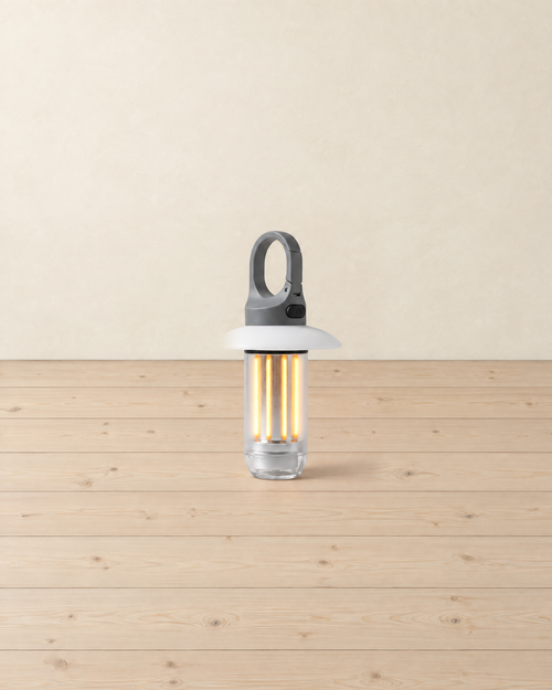 Small camping lantern with a grey clip hook, white shade and clear tube of four warm LED strips, standing on pale pine floorboards