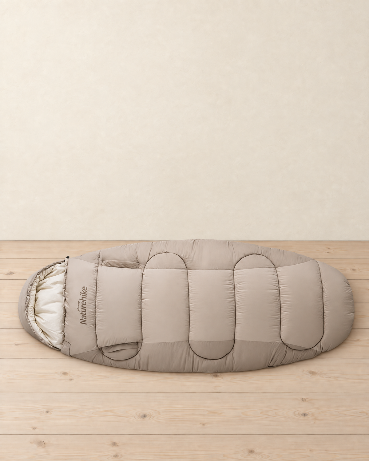 Light beige wide hooded sleeping bag with arm openings laid flat on a pale pine floor, hood with its cream lining at the left