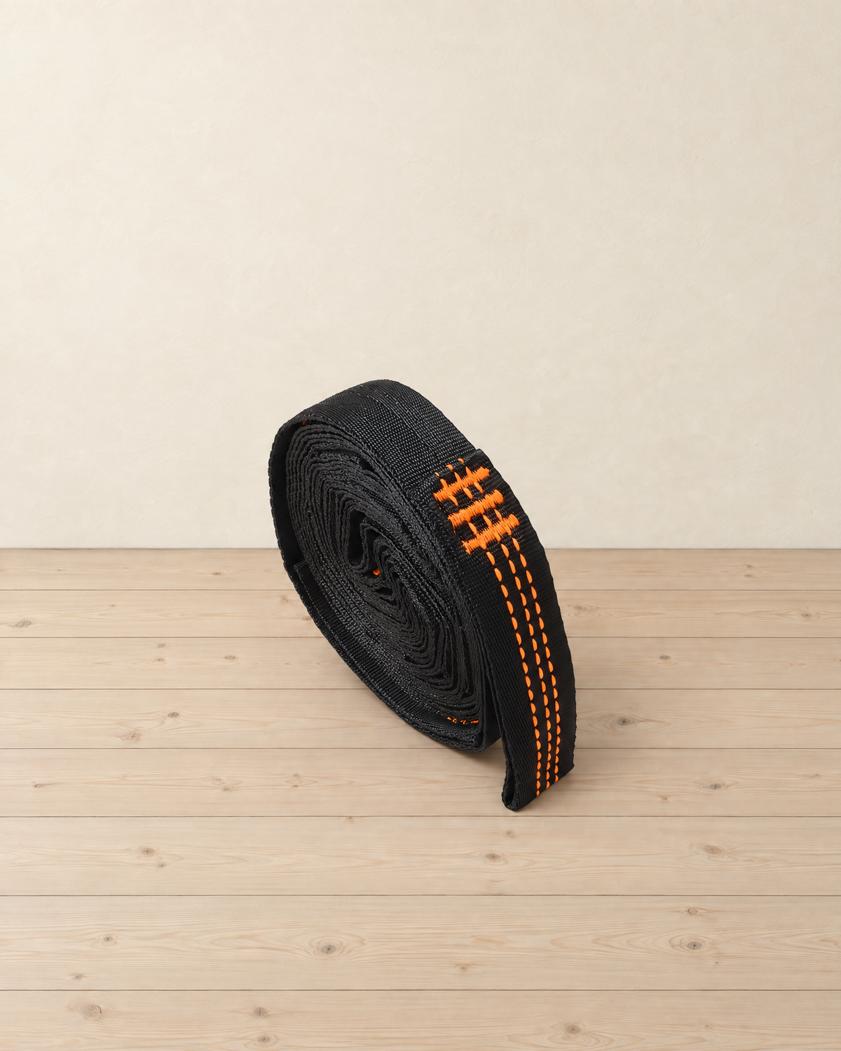 Black hammock tree strap with orange stitching rolled into a tight coil on pale pine floorboards