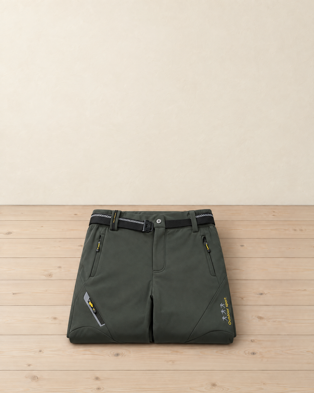 Dark green men's fleece-lined softshell hiking trousers with a black belt, folded on a pale pine floor, showing the zip pockets and the yellow thigh lettering