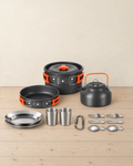 Black and orange camping cook set for two on a pale pine floor: pot with lid, frying pan, kettle, two steel plates, two cups, a carabiner and six folding cutlery pieces