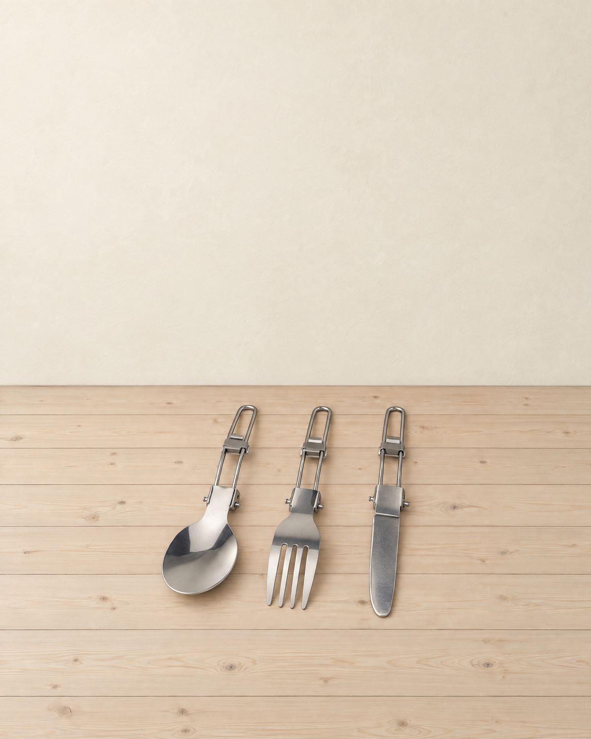 Folding stainless steel spoon, fork and table knife lying unfolded side by side on a pale pine floor, each with a wire loop handle