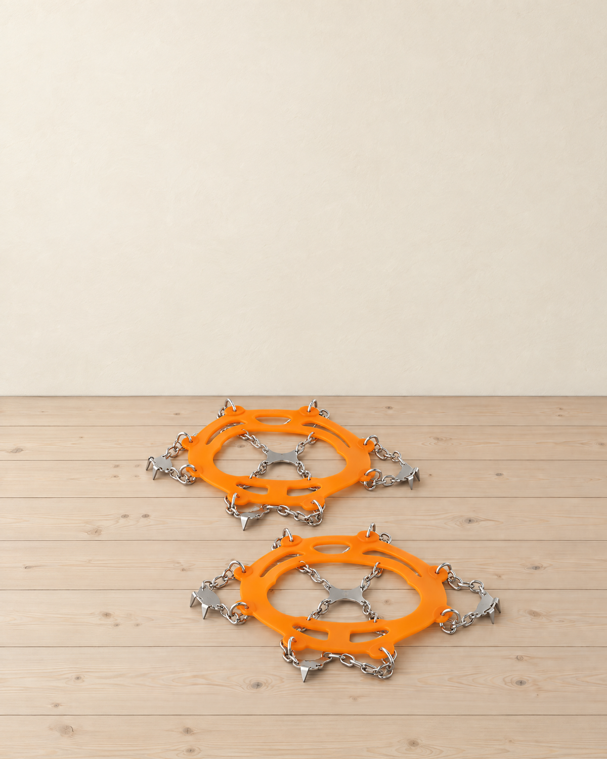 Pair of orange chain crampons lying flat on a pale pine floor, with the stretch harness, steel chains and toothed steel plates