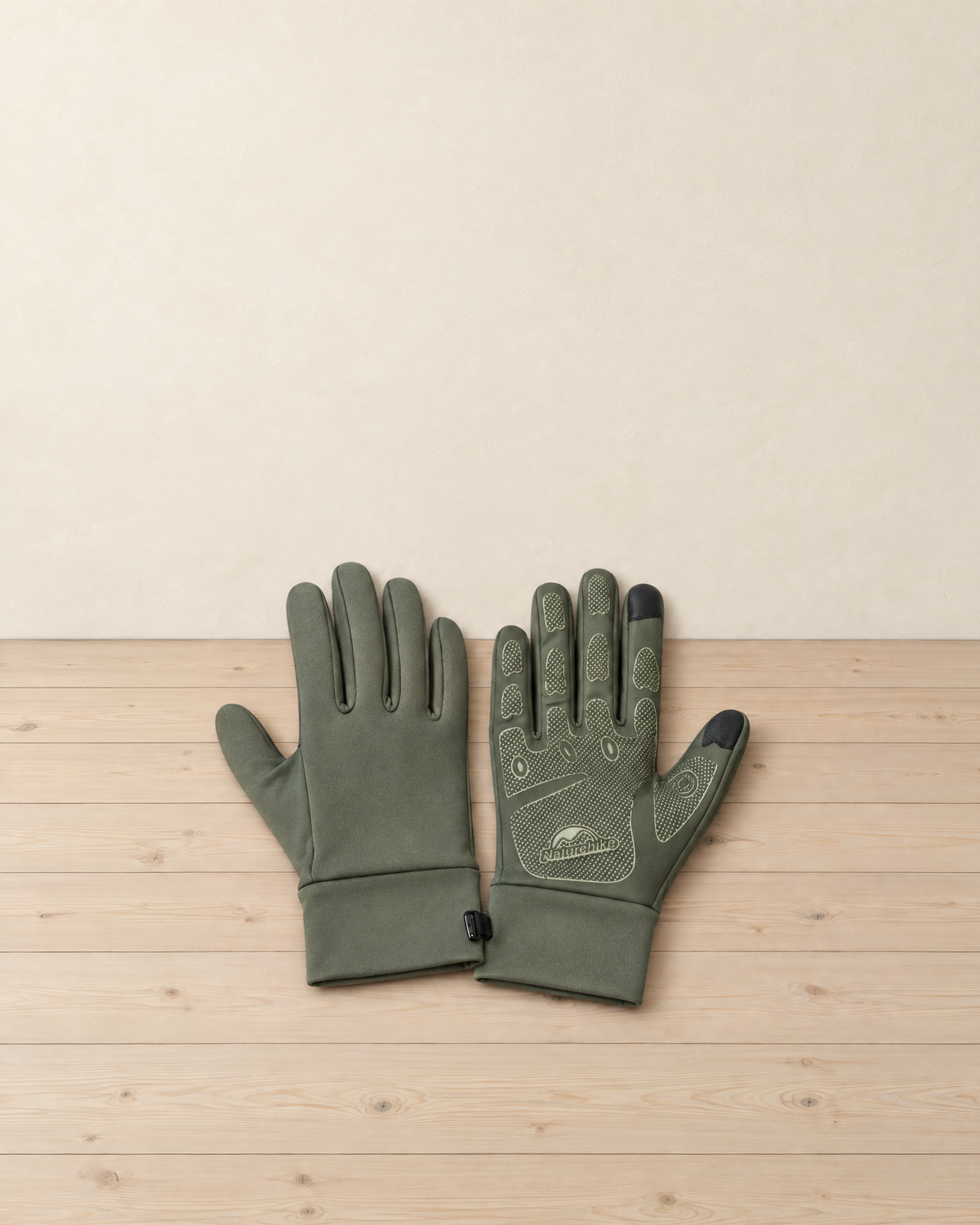 Pair of army green stretch hiking gloves on a pale pine floor, one back up and one palm up with its dotted grip and touchscreen fingertips
