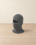 Grey rib-knit winter balaclava with one wide eye opening standing on pale pine floorboards, its neck flared at the base