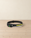 Black LED headlamp with a yellow light strip, round spotlight and two-button control module, standing on pale pine floorboards against a cream wall