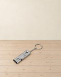 Silver grey double-tube metal whistle on a short chain and split ring, lying on pale pine floorboards