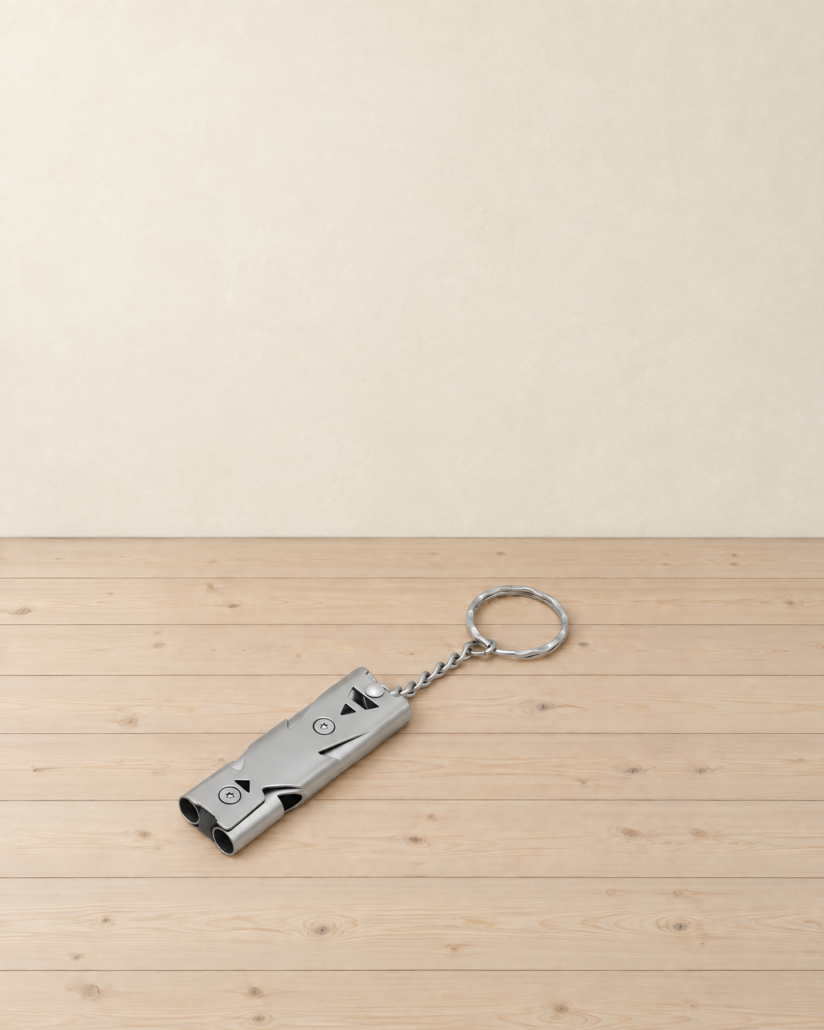 Silver grey double-tube metal whistle on a short chain and split ring, lying on pale pine floorboards