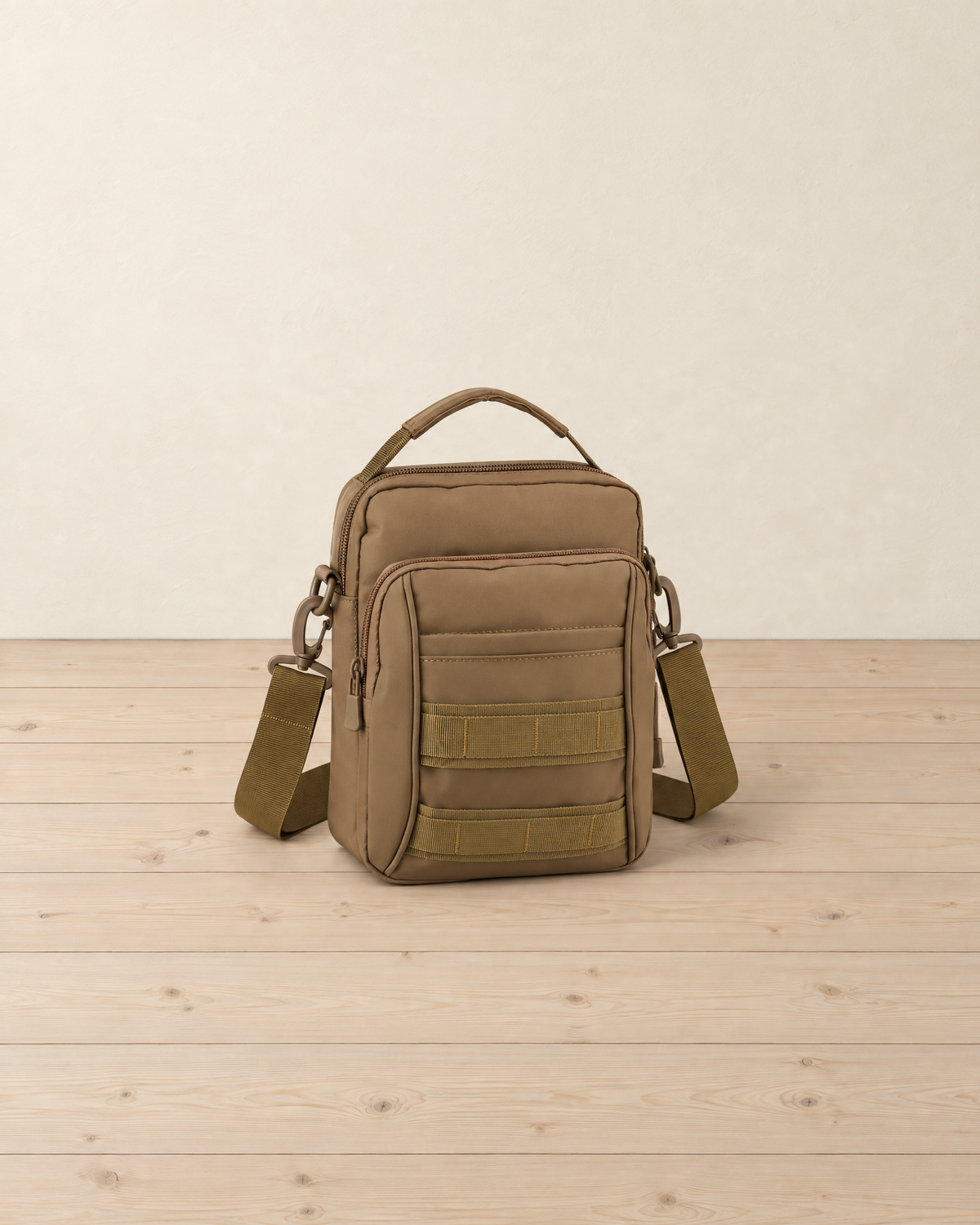 Coyote brown tactical shoulder bag standing on a pale pine floor, with its carry handle, zipped front pocket, two webbing bands and clipped-on strap