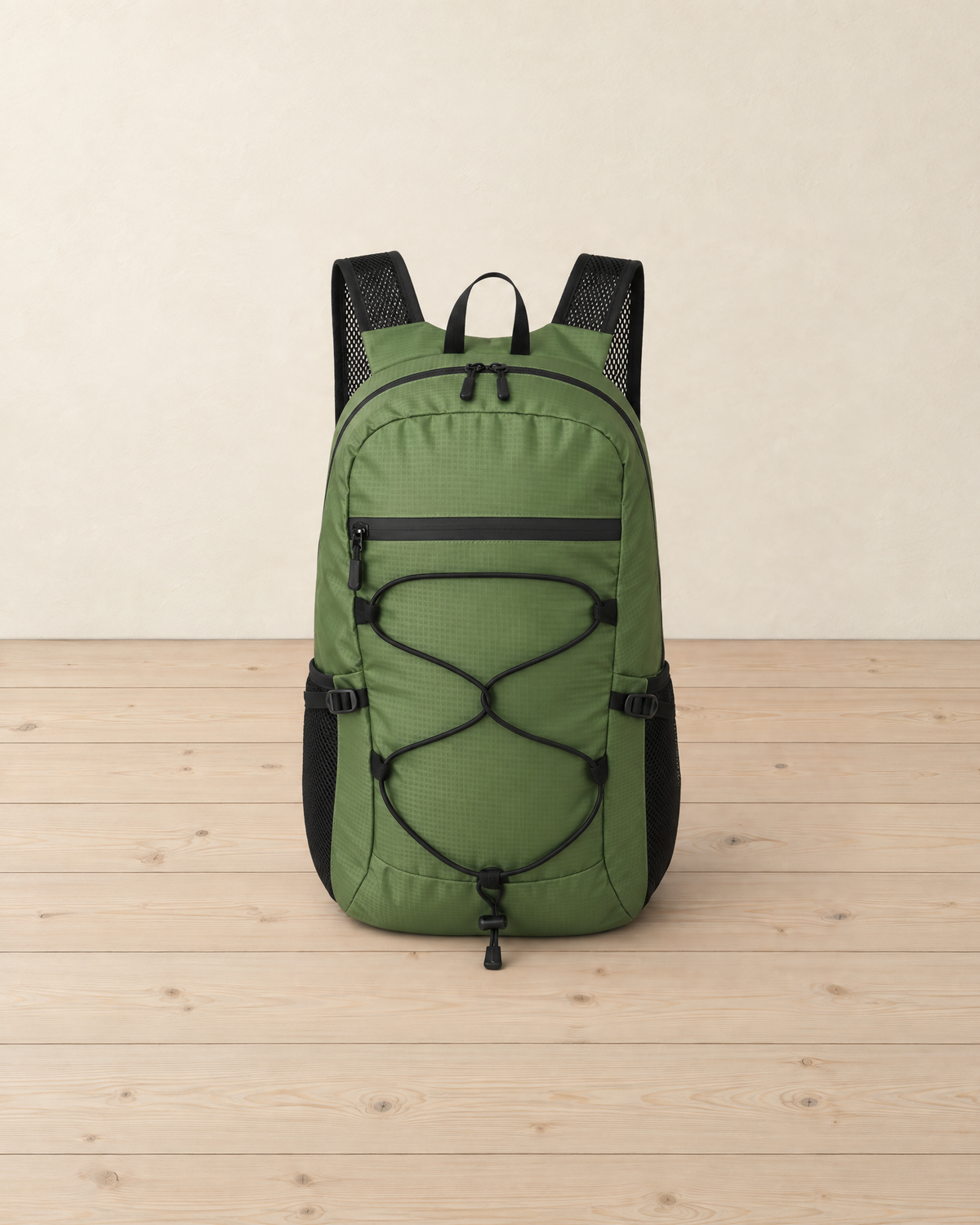 Army green packable daypack with a black criss-cross bungee cord, front zip pocket and black mesh side pockets, standing on pale pine floorboards against a cream wall