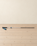 Silver and black folding trekking pole extended and lying on pale pine floorboards, its black foam grip and blue wrist strap at the left and the blue lock collar on the silver shaft