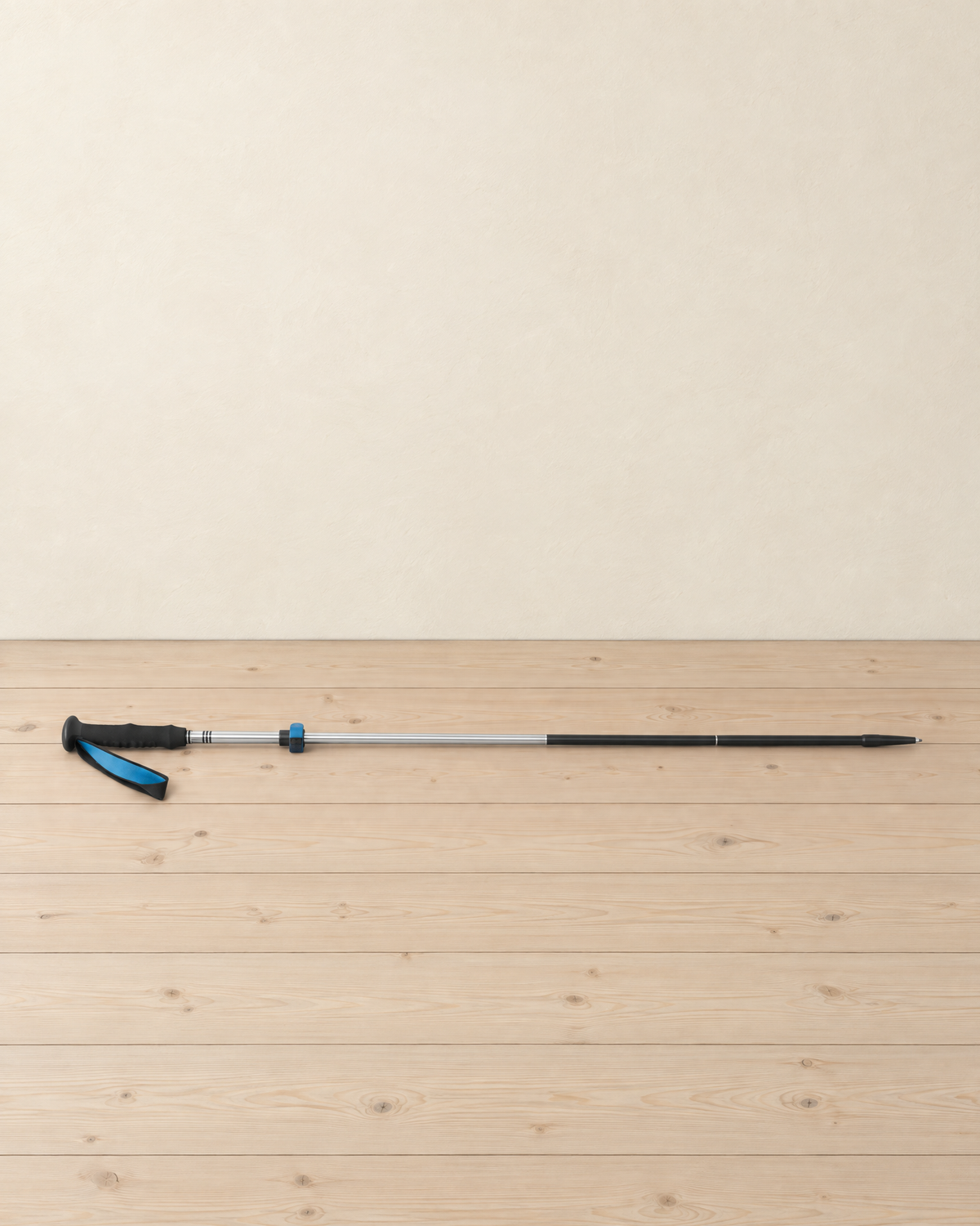 Silver and black folding trekking pole extended and lying on pale pine floorboards, its black foam grip and blue wrist strap at the left and the blue lock collar on the silver shaft