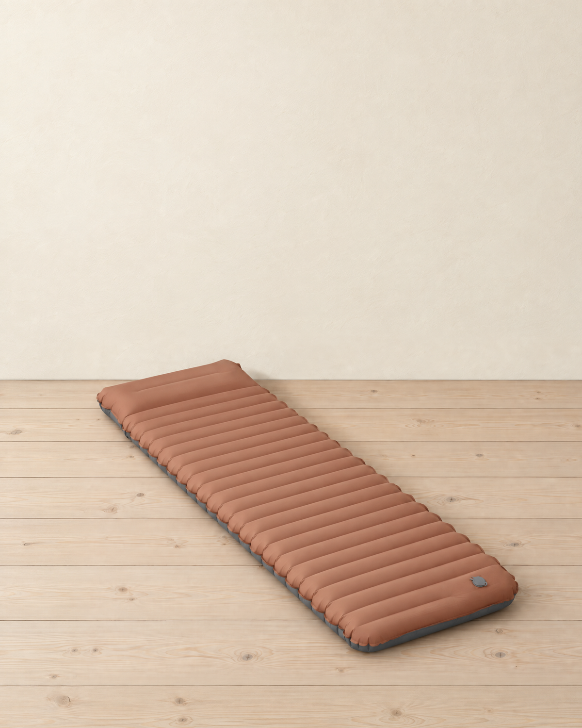 Brown inflatable sleeping mat with grey underside lying on a pale pine floor, showing its cross tubes, raised head section and grey valve