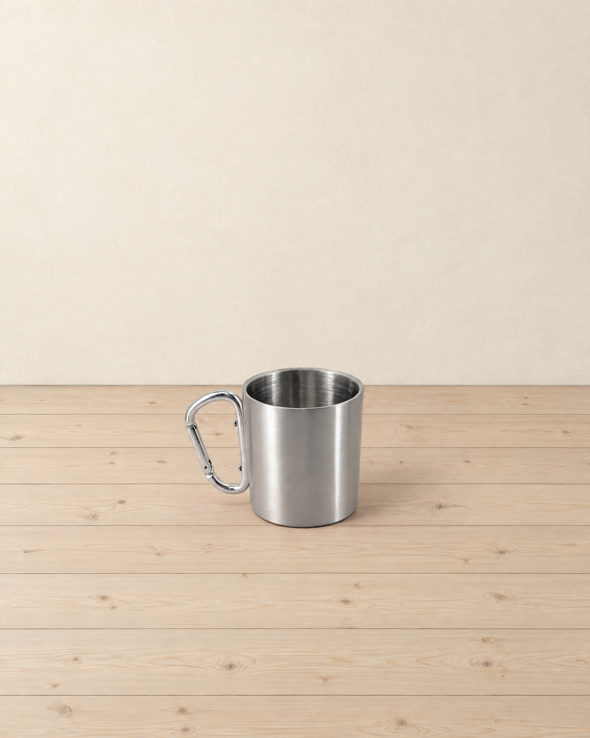 Stainless steel camping mug with a carabiner handle standing on pale pine floorboards against a cream wall