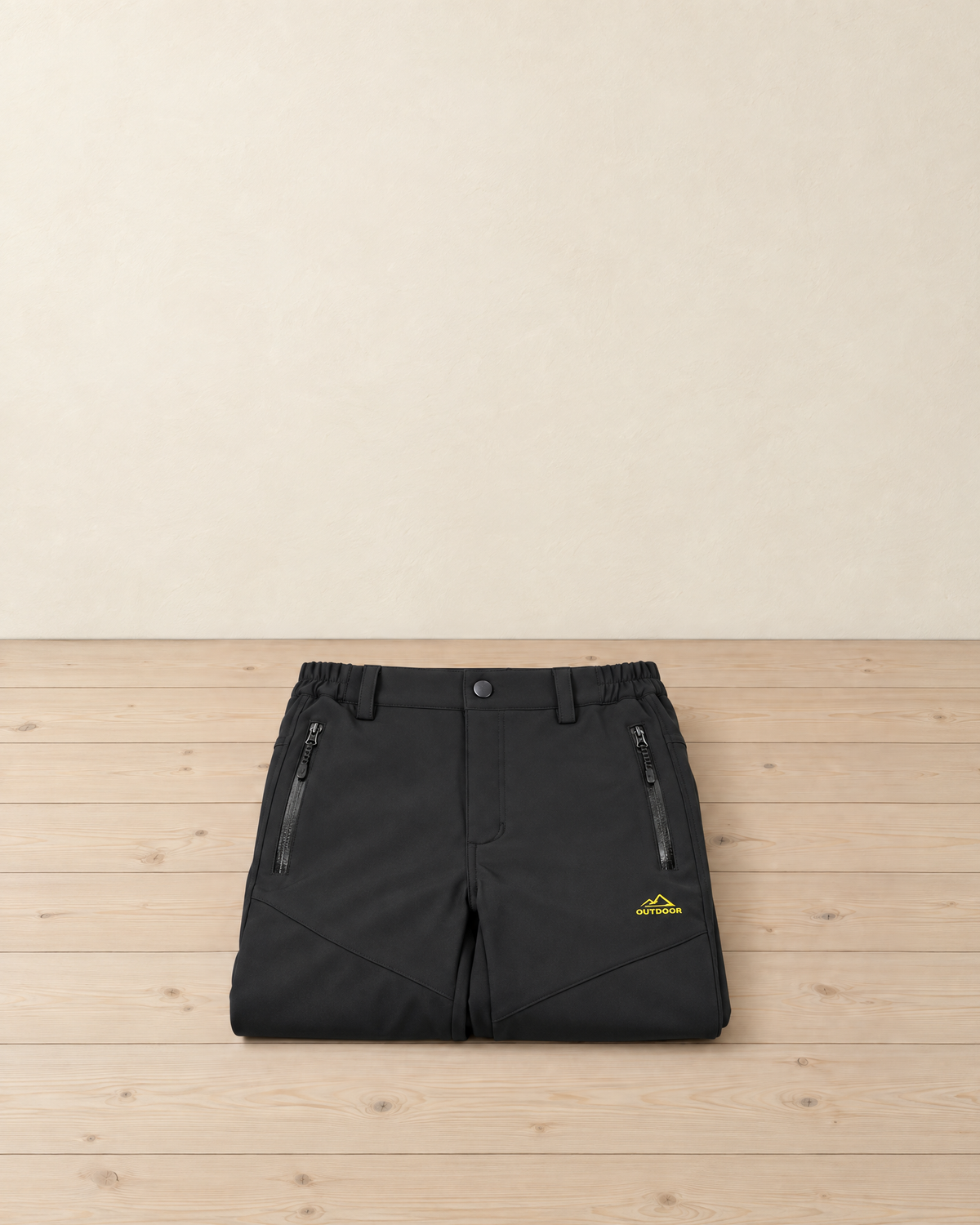 Black women's fleece-lined softshell hiking trousers folded on a pale pine floor, showing the snap waistband, two zip pockets and the small yellow thigh embroidery