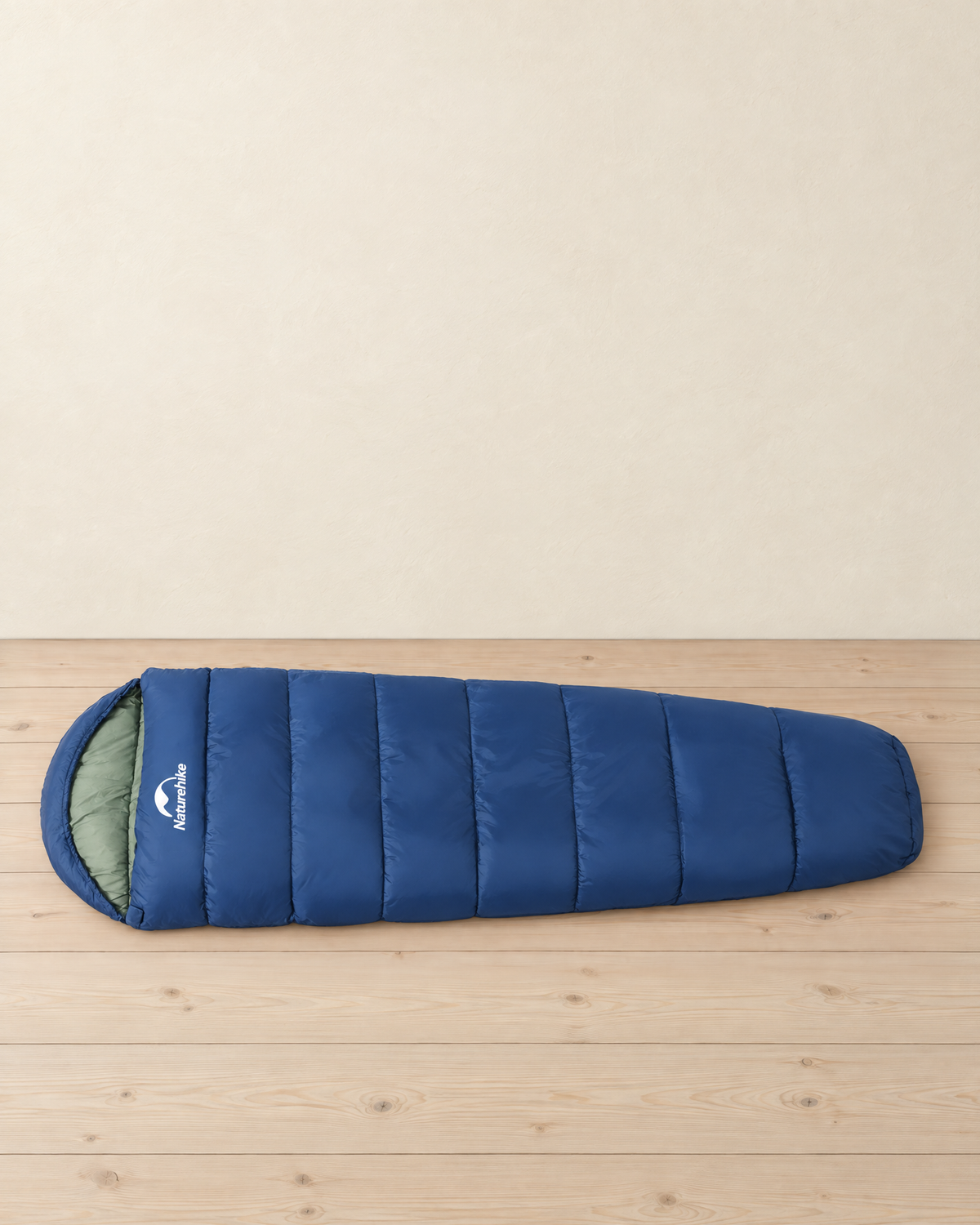 Navy blue mummy sleeping bag with a light green lining laid flat on a pale pine floor, hood at the left