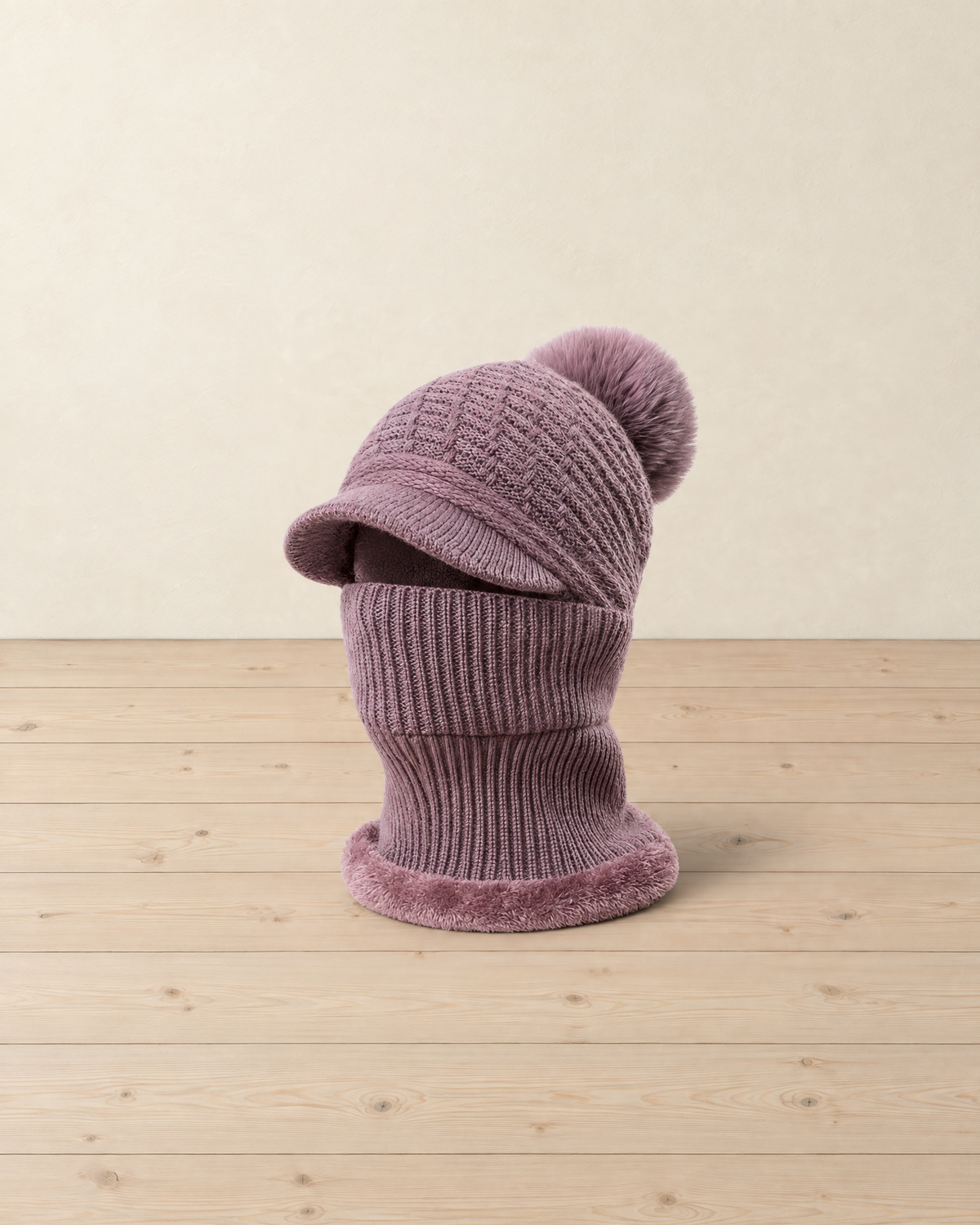Dusty mauve knitted brim beanie with a faux-fur pompom and a ribbed neck warmer with a plush edge, on pale pine floorboards