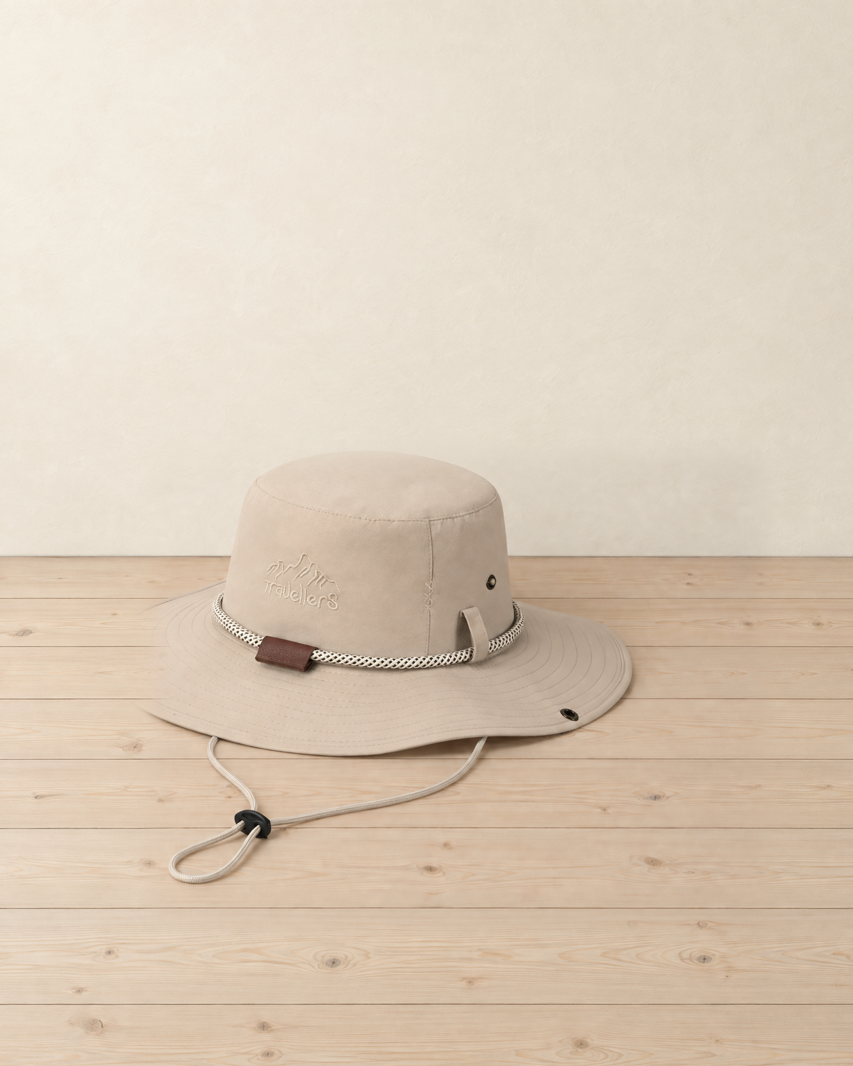 Beige wide brim hiking hat with a braided cord band, brown tab and chin strap, resting on a pale pine floor