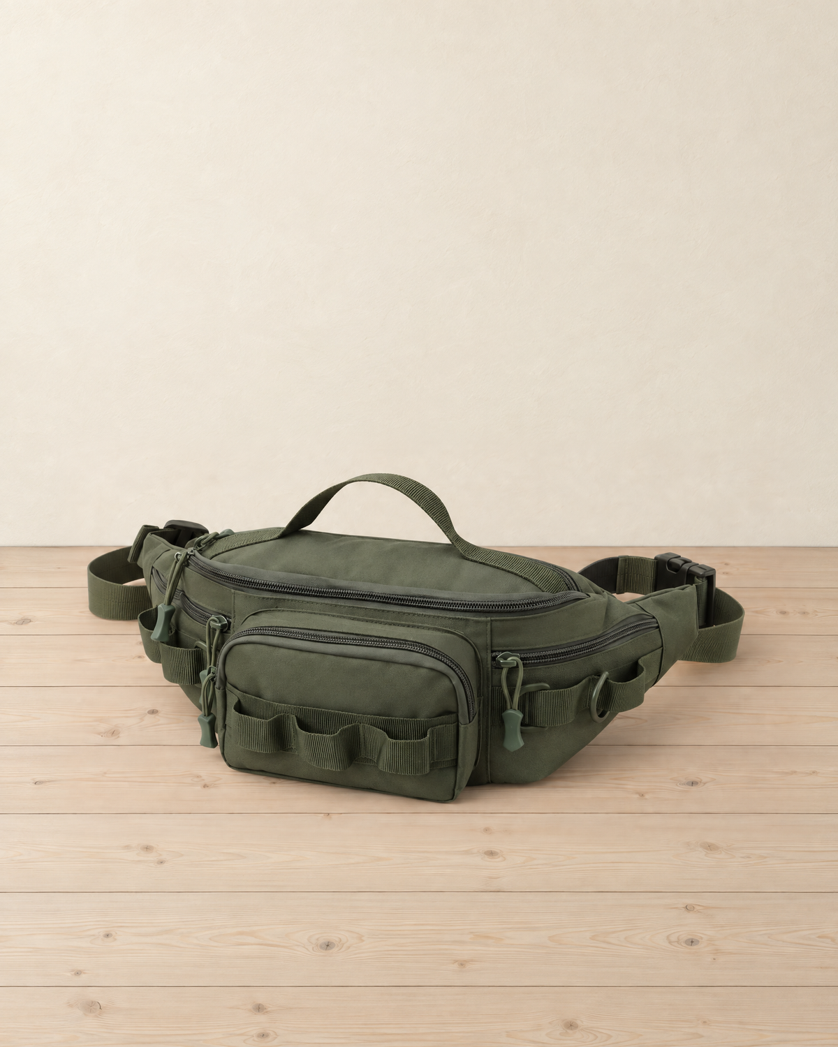 Army green hip pack with a top grab handle and front gear loops standing on pale pine floorboards against a cream wall