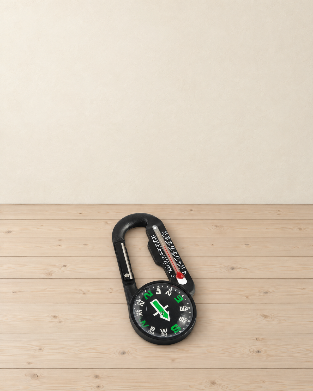 Black compass carabiner with a thermometer and a green-marked compass lying on a pale pine floor