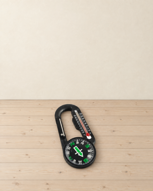 Black compass carabiner with a thermometer and a green-marked compass lying on a pale pine floor