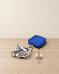 Stainless steel camping stove burner with four folding pot supports, its brass valve stem and a blue zipped pouch on a pale pine floor