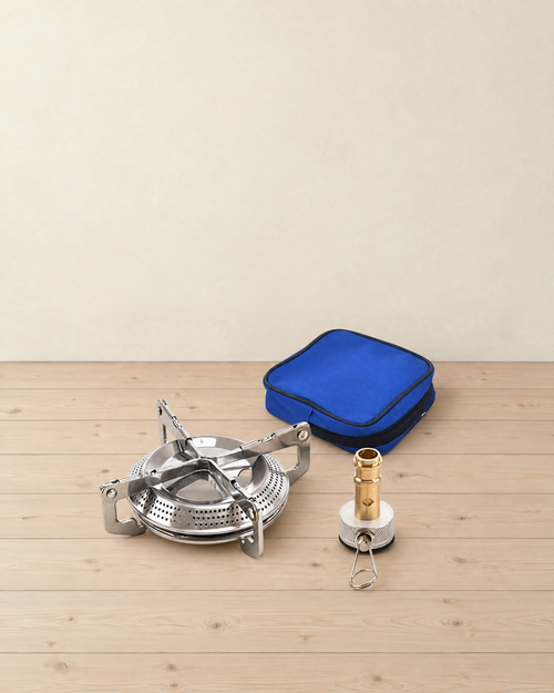 Stainless steel camping stove burner with four folding pot supports, its brass valve stem and a blue zipped pouch on a pale pine floor