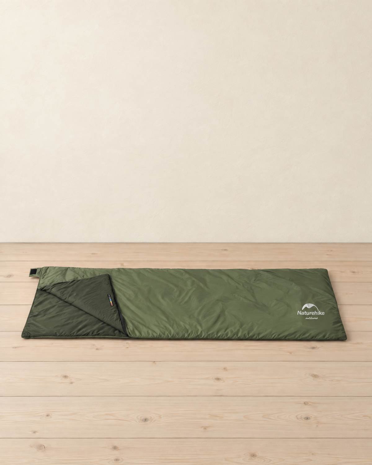 Army green envelope sleeping bag laid flat on pale pine floorboards, its head corner folded back to show the dark green lining and the white logo printed at the foot end