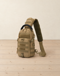 Sand beige tactical sling bag with webbing loops and a black braided handle standing on pale pine floorboards