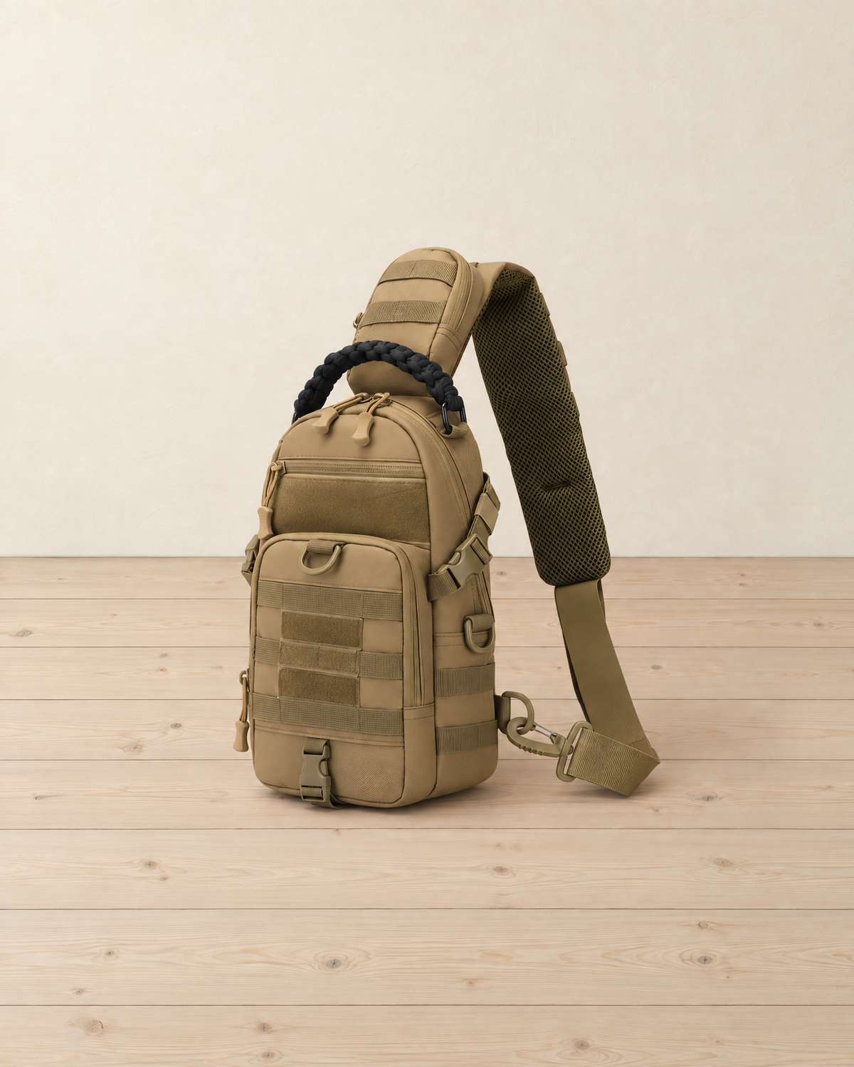 Sand beige tactical sling bag with webbing loops and a black braided handle standing on pale pine floorboards