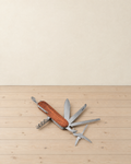 Wooden-handled pocket multitool with its blade, two saw blades, scissors, opener and corkscrew folded out on a pale pine floor
