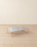 Brushed stainless steel folding mini camping table with thin wire legs, standing on pale pine floorboards against a cream plaster wall