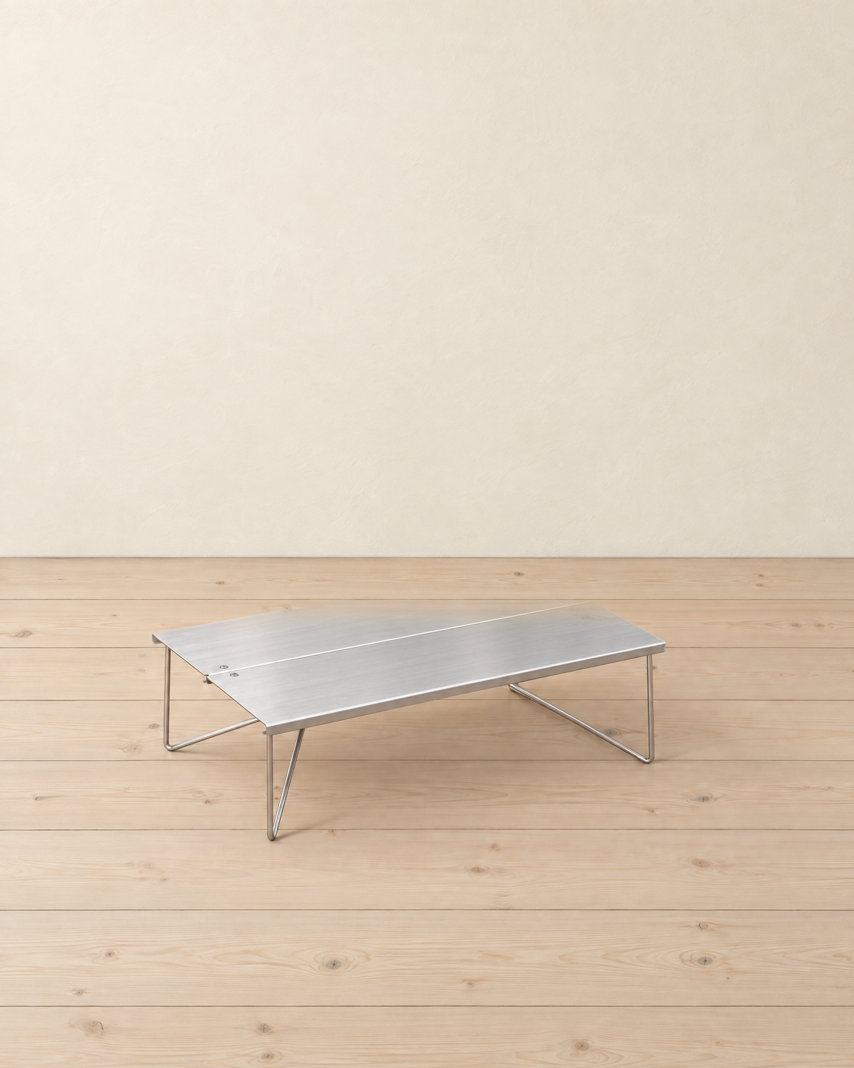 Brushed stainless steel folding mini camping table with thin wire legs, standing on pale pine floorboards against a cream plaster wall