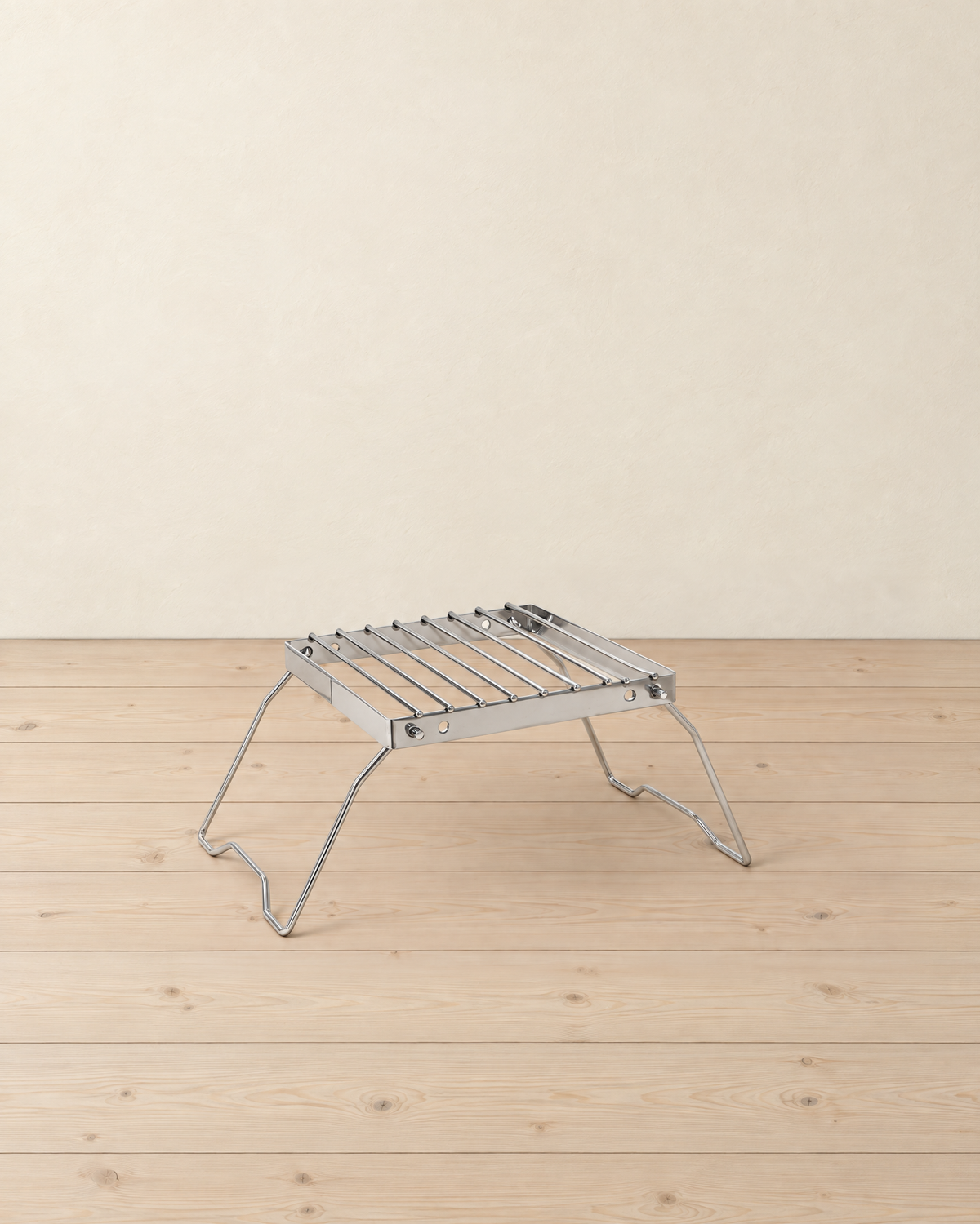 Folding stainless steel grill stand with a nine-bar grate on two splayed wire legs, on a pale pine floor against a cream wall