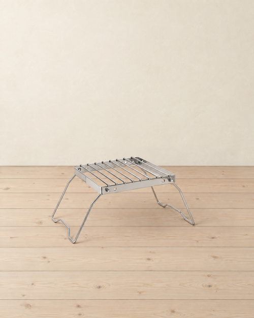 Folding stainless steel grill stand with a nine-bar grate on two splayed wire legs, on a pale pine floor against a cream wall