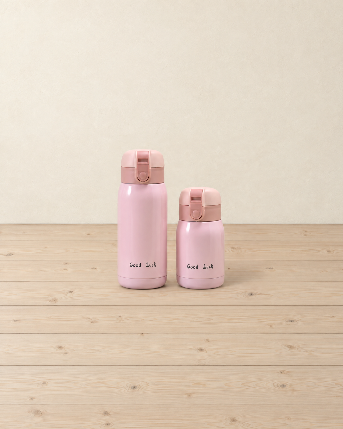 Pink mini insulated bottles in 360 ml and 200 ml with flip lids, printed Good Luck, standing on a pale pine floor