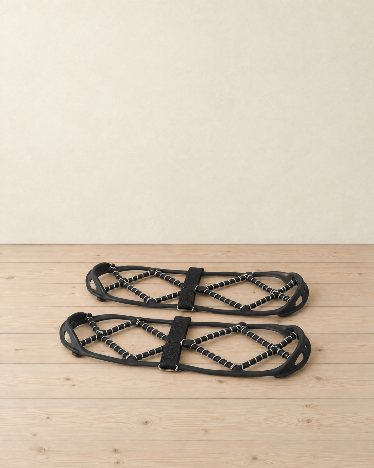 Pair of black ice grips with steel coil cords and instep straps lying on a pale pine floor
