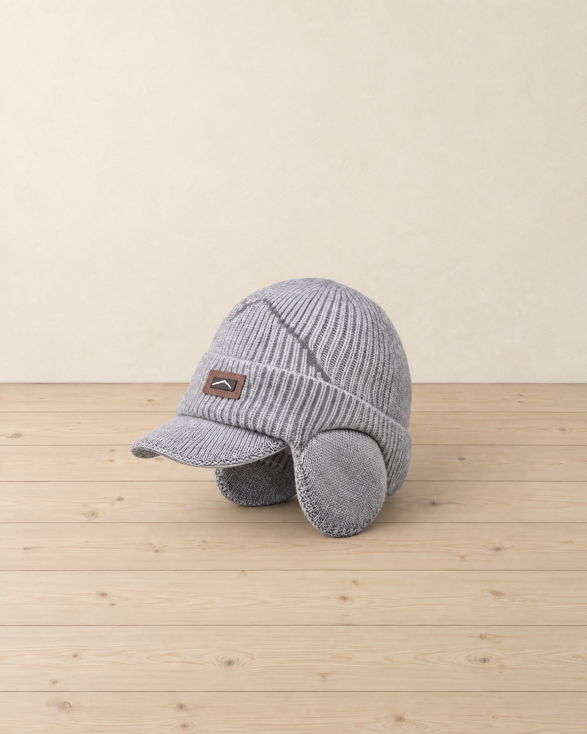 Light grey ribbed knit peaked beanie with round ear flaps, a zigzag line across the crown and a small leather patch, on pale pine floorboards
