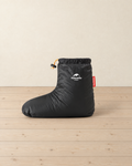 Black down bootie standing in side profile on pale pine floorboards, with its gathered elastic cuff, orange drawcord and white printed logo