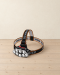Black rechargeable headlamp with eight round LEDs on a black and orange head strap printed LED HEADLIGHT, on a pale pine floor