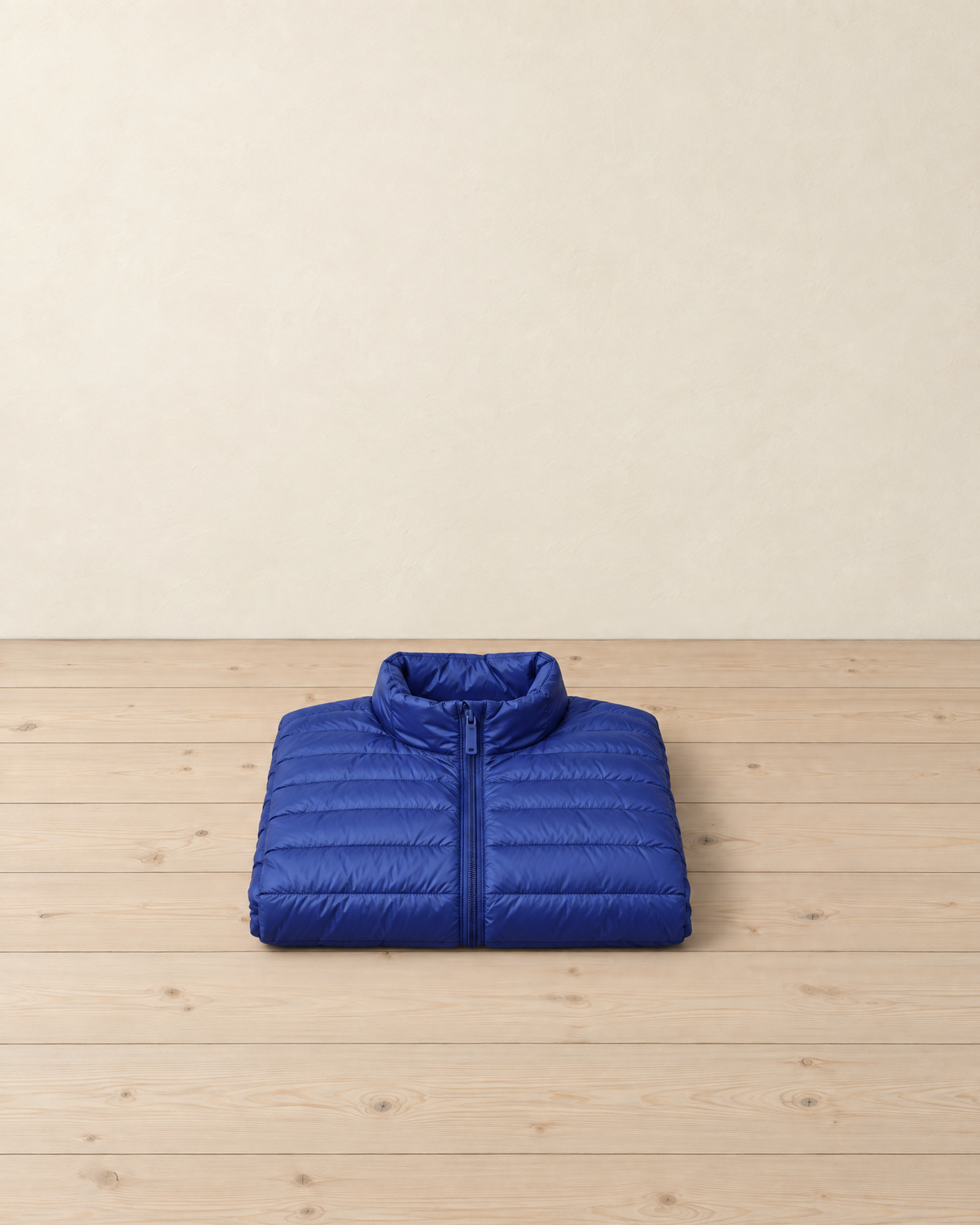 Royal blue men's lightweight puffer jacket folded on a pale pine floor, showing the low stand collar, centre zip and narrow horizontal quilting