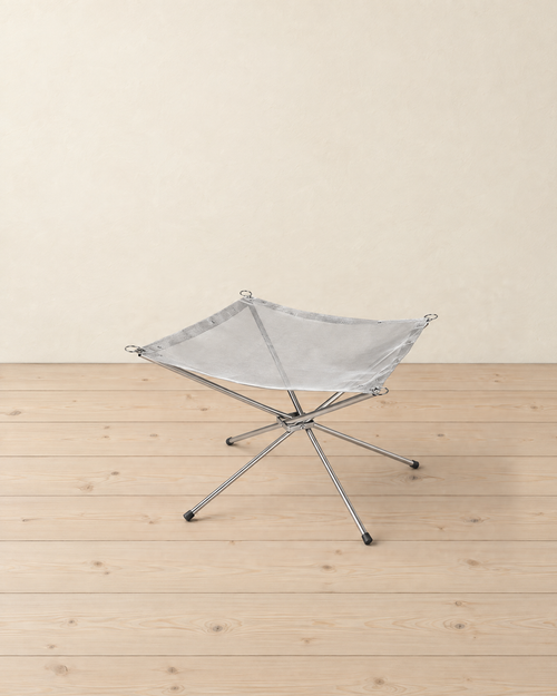 Empty folding fire pit on a pale pine floor: a square stainless steel mesh with corner rings on four crossed steel legs with rubber feet