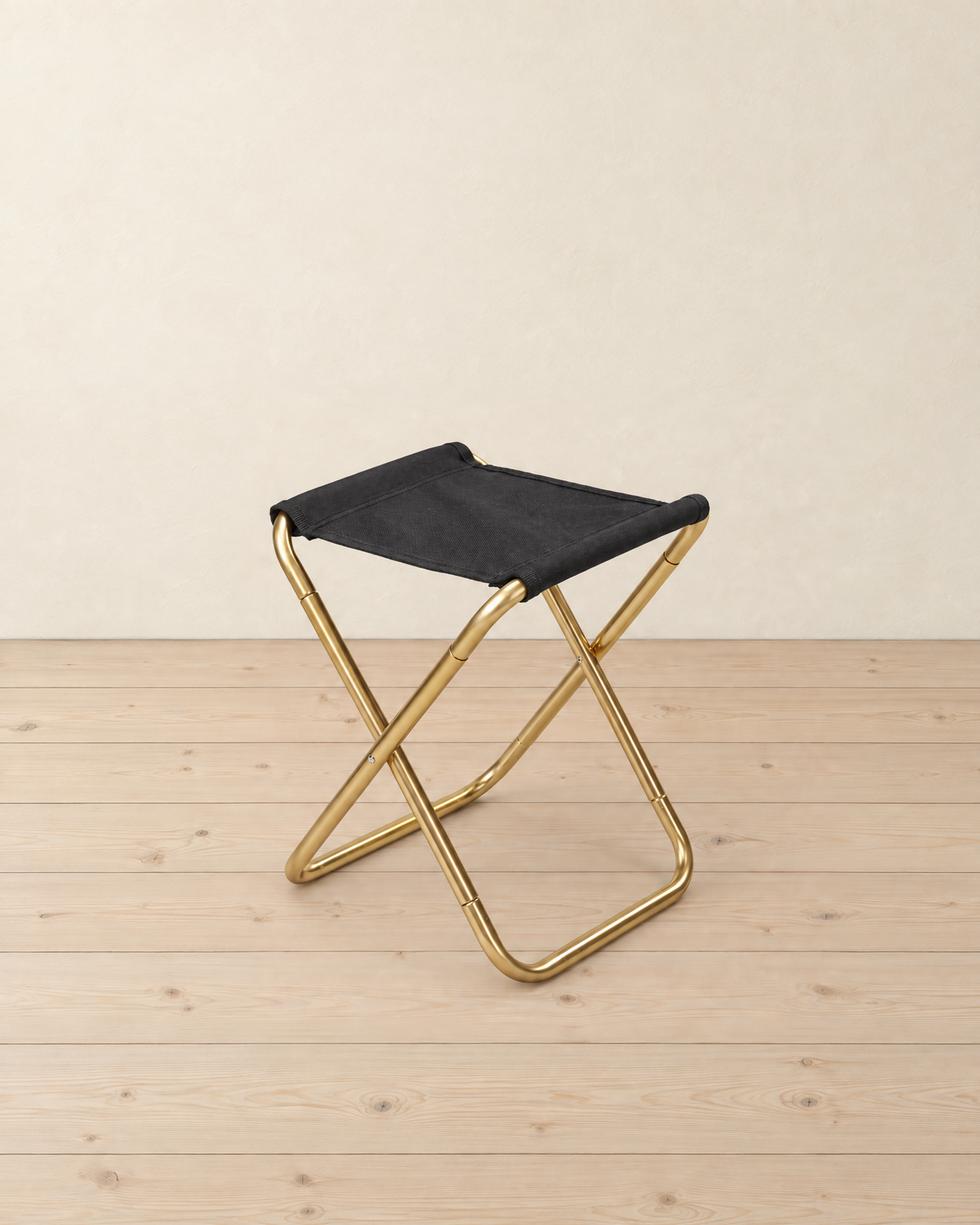 Gold aluminium folding camp stool with a black fabric seat, standing on pale pine floorboards against a cream plaster wall