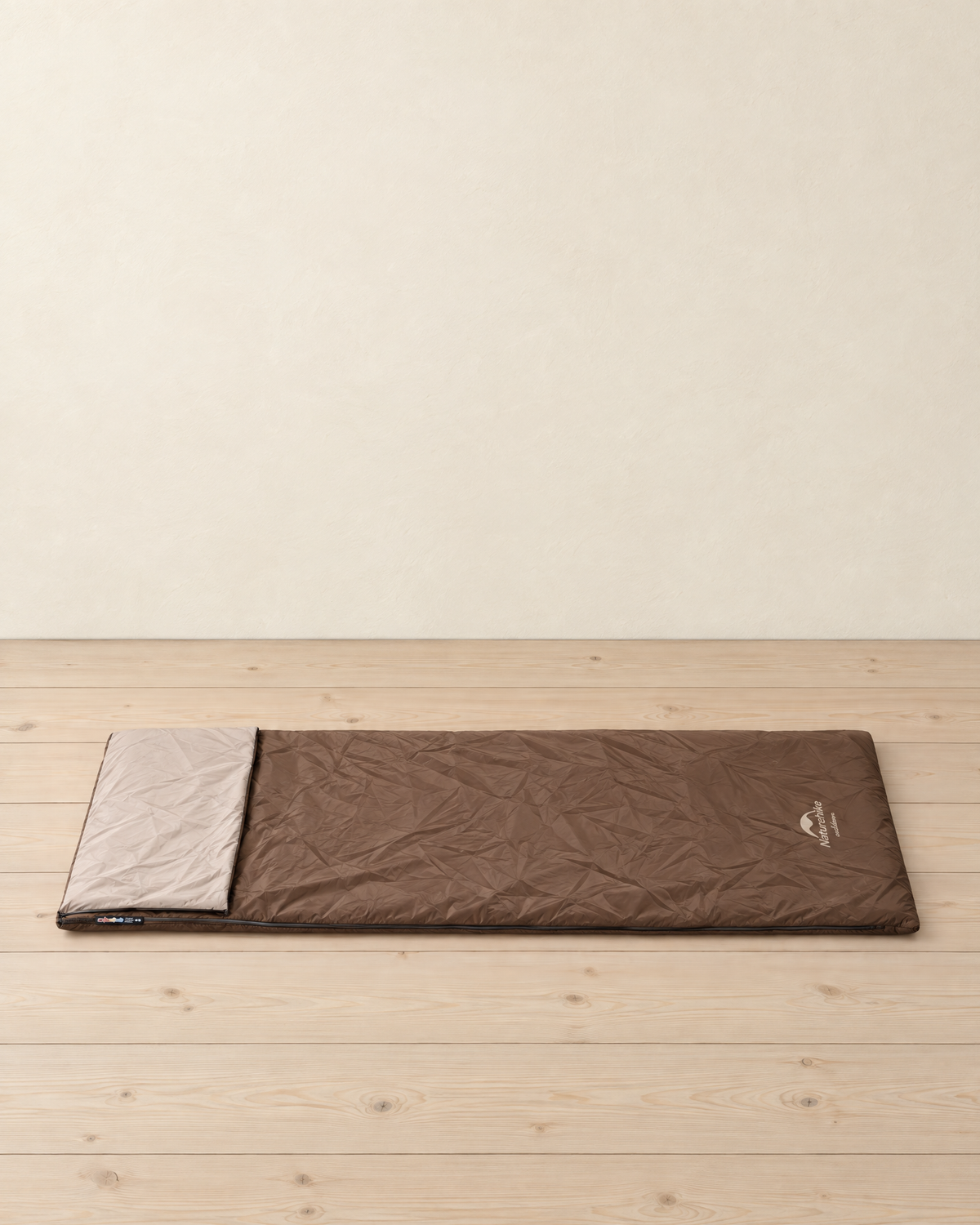 Brown envelope sleeping bag lying flat on a pale pine floor, its head folded back to show the beige lining, the maker's logo at the foot