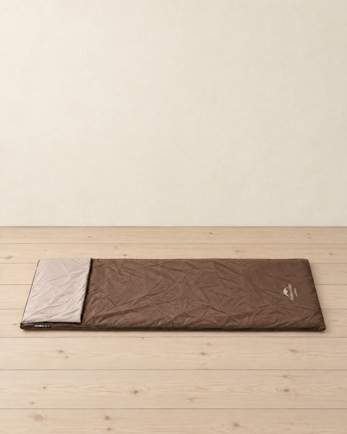 Brown envelope sleeping bag lying flat on a pale pine floor, its head folded back to show the beige lining, the maker's logo at the foot