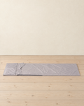 Light grey sleeping bag liner laid out flat on pale pine floorboards, the pillow flap at the head and a zip along one side