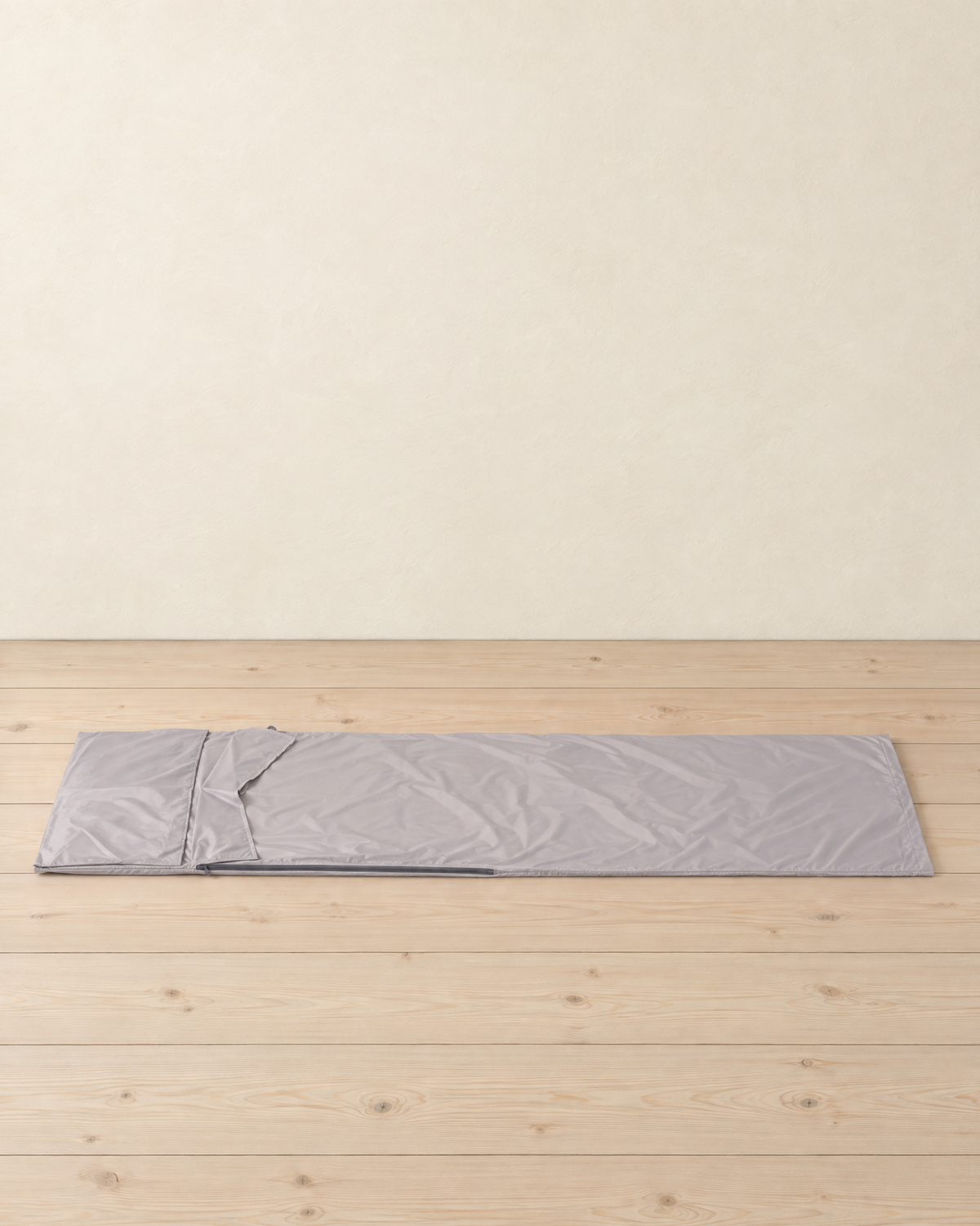 Light grey sleeping bag liner laid out flat on pale pine floorboards, the pillow flap at the head and a zip along one side