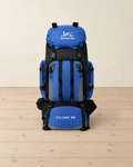 Blue and black 90 L hiking backpack standing upright on a pale pine floor, lid pocket, front pocket straps and side pockets facing the camera