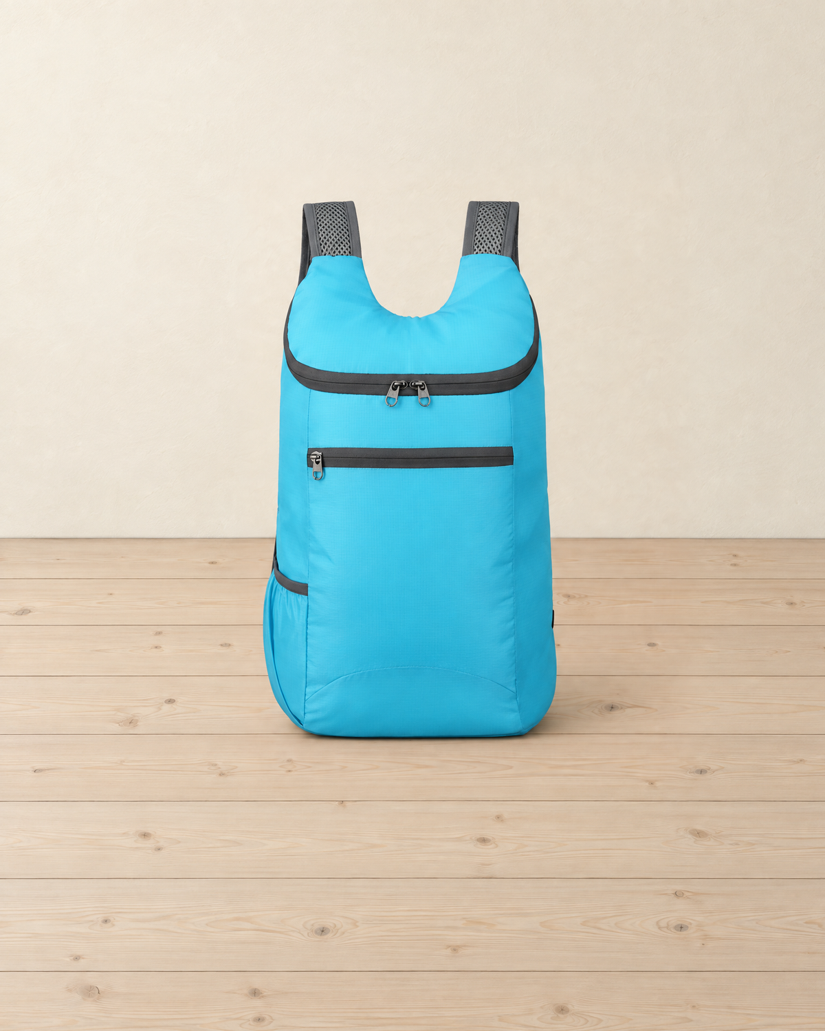 Sky blue packable daypack standing on a pale pine floor, with a curved top zip, a zipped front pocket and grey mesh shoulder straps