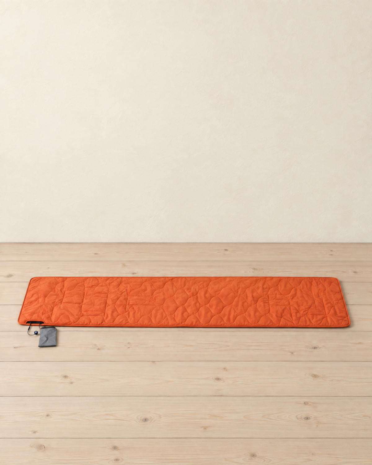 Bright orange quilted USB heated sleeping pad lying flat on pale pine floorboards, its power bank pouch beside it